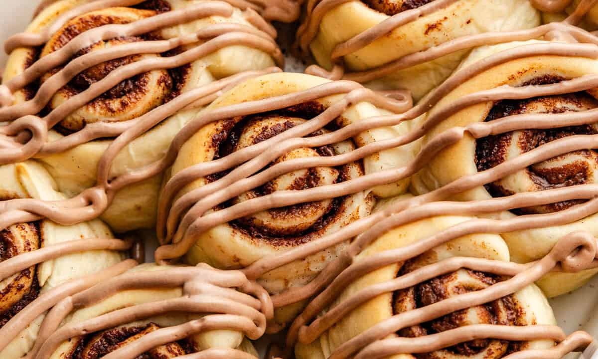 A close up view of nutella cinnamon rolls in a pan, topped with a drizzle of nutella frosting.