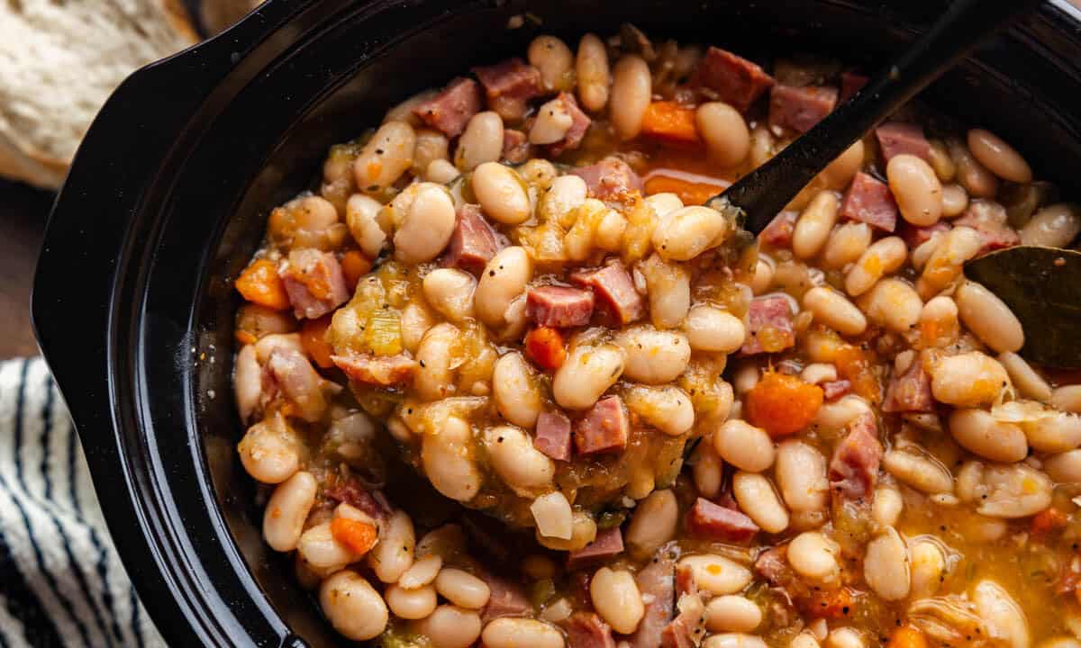 An overhead view of a slow cooker of ham and beans with a ladle dishing out a scoop.