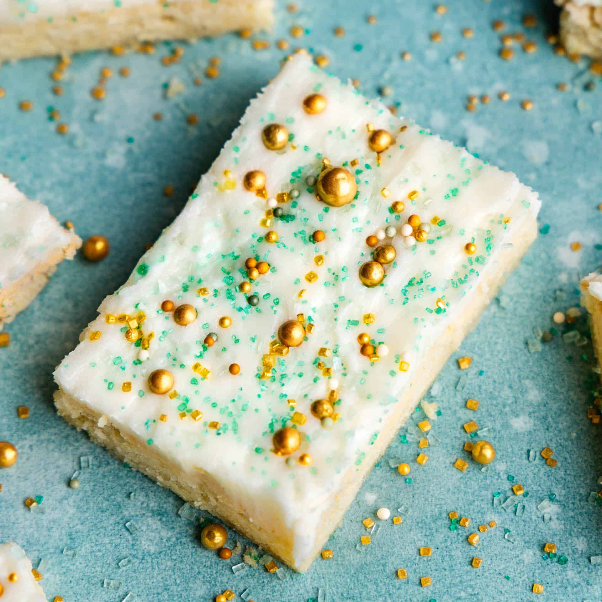 A sugar cookie bar on a blue background topped with blue and gold sprinkles and frosted with a white frosting.