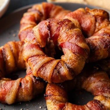 A plate of piled bacon wrapped sausage filled onion rings.