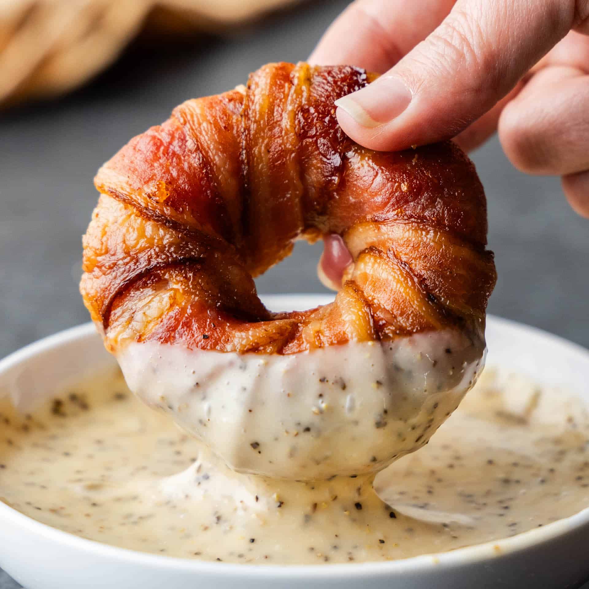 A bacon wrapped sausage filled onion ring being dipped in white sauce.