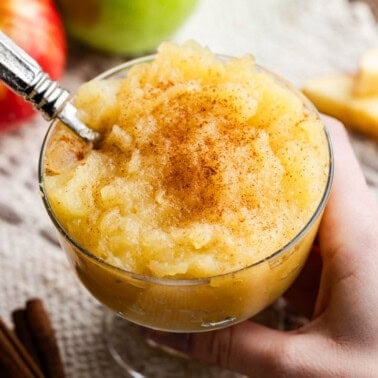 Fancy glass full of applesauce with a spoon in it being held.