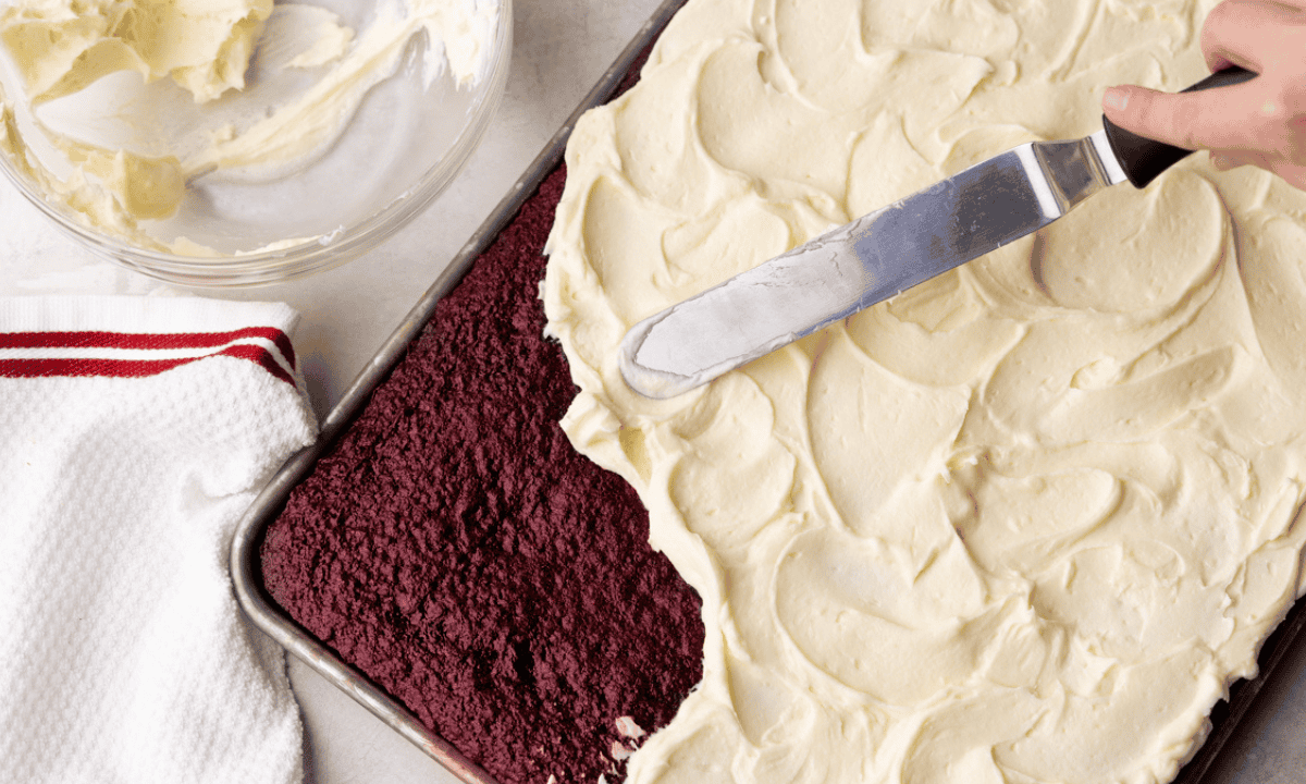 An overhead view of a red velvet sheet cake that is being frosted.