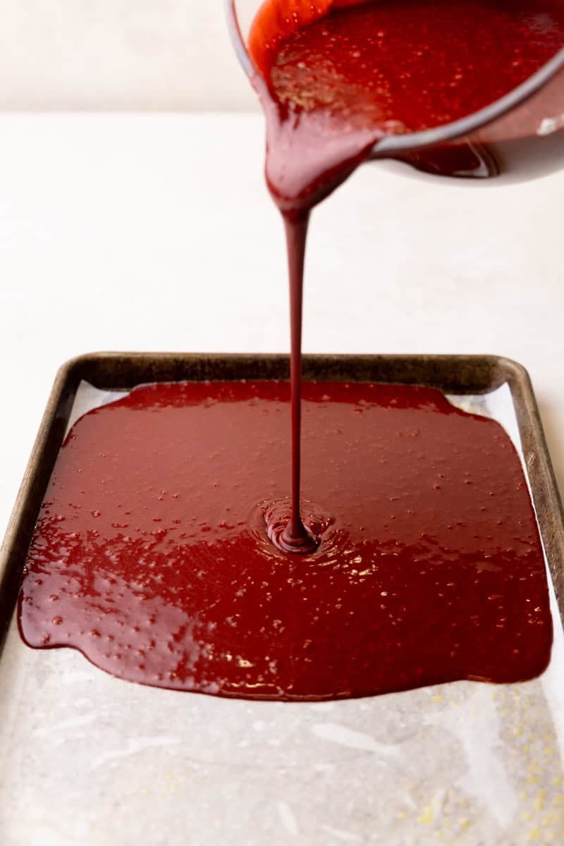 Red velvet cake batter being poured from a mixing bowl onto a parchment lined baking sheet.