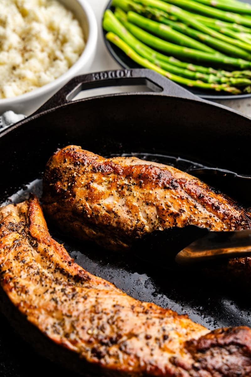 Closeup of tongs picking up Garlic Butter Pork Tenderloin on a skillet with mashed potatoes and asparagus in the background.