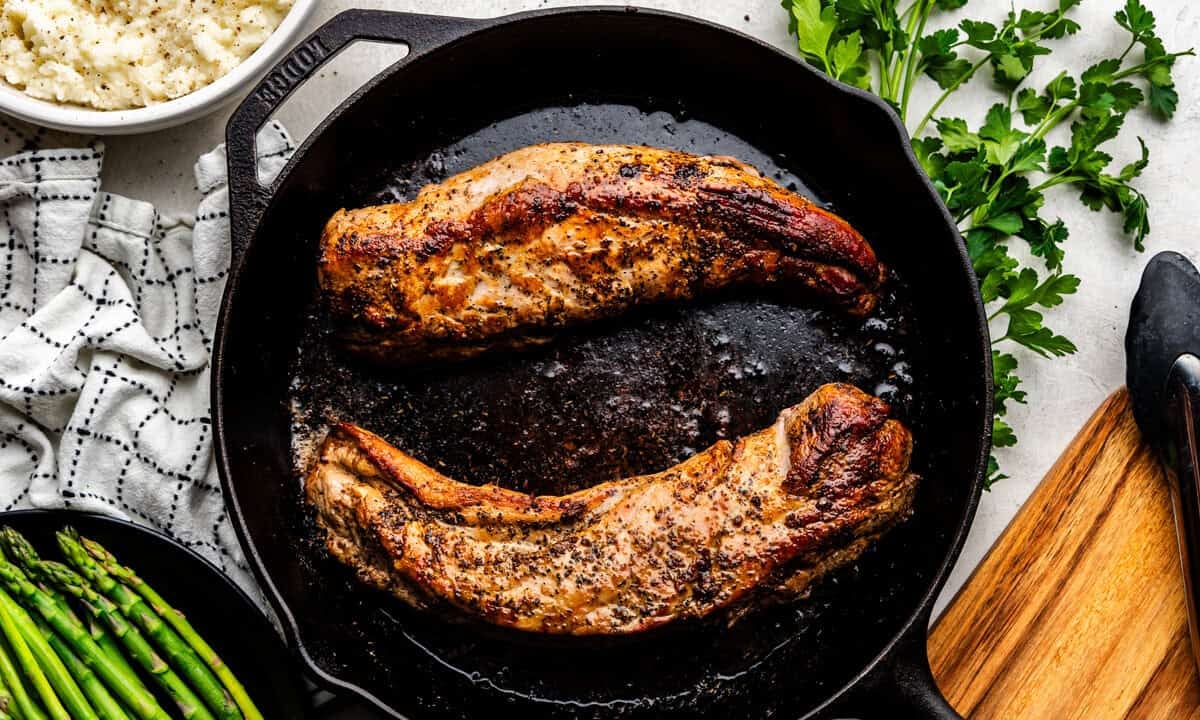 Overhead of Garlic Butter Pork Tenderloin on a skillet with mashed potatoes and asparagus in the background.