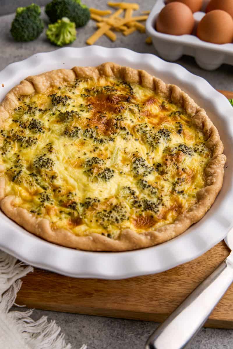 A whole Ham, Broccoli & Cheddar Quiche with ingredients and eggs in the background.