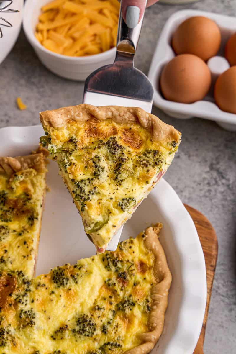 A slice of Ham, Broccoli & Cheddar Quiche with ingredients and eggs in the background.