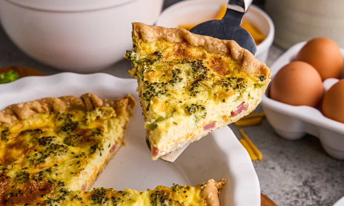 A slice of Ham, Broccoli & Cheddar Quiche with ingredients and eggs in the background.