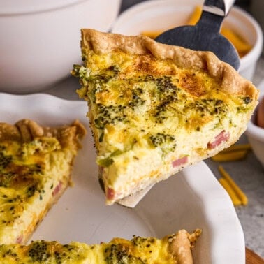 A slice of Ham, Broccoli & Cheddar Quiche with ingredients and eggs in the background.