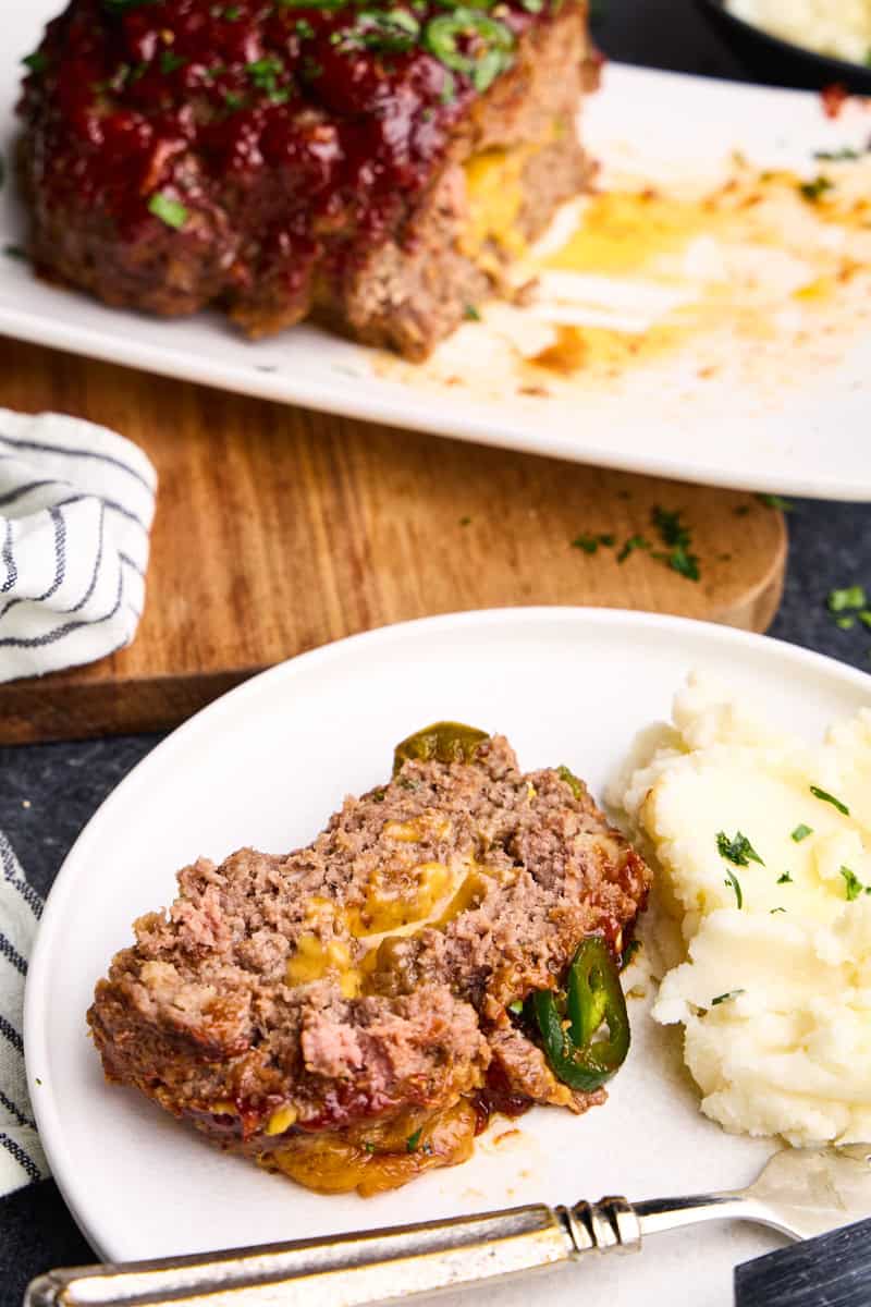 Slice of Jalapeno Cheddar Stuffed Meatloaf on a plate with mashed potatoes and meatloaf in the background.