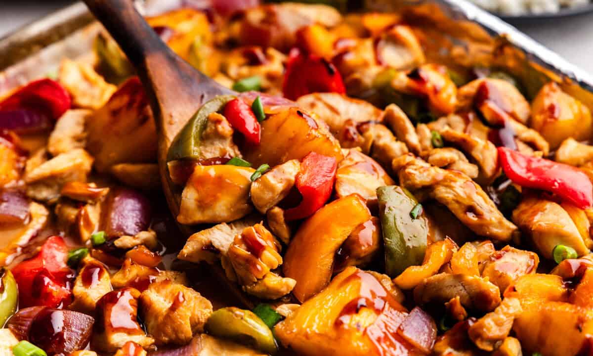 Closeup of Hawaiian Chicken & Veggies on a sheet pan lifted by a wooden spoon.