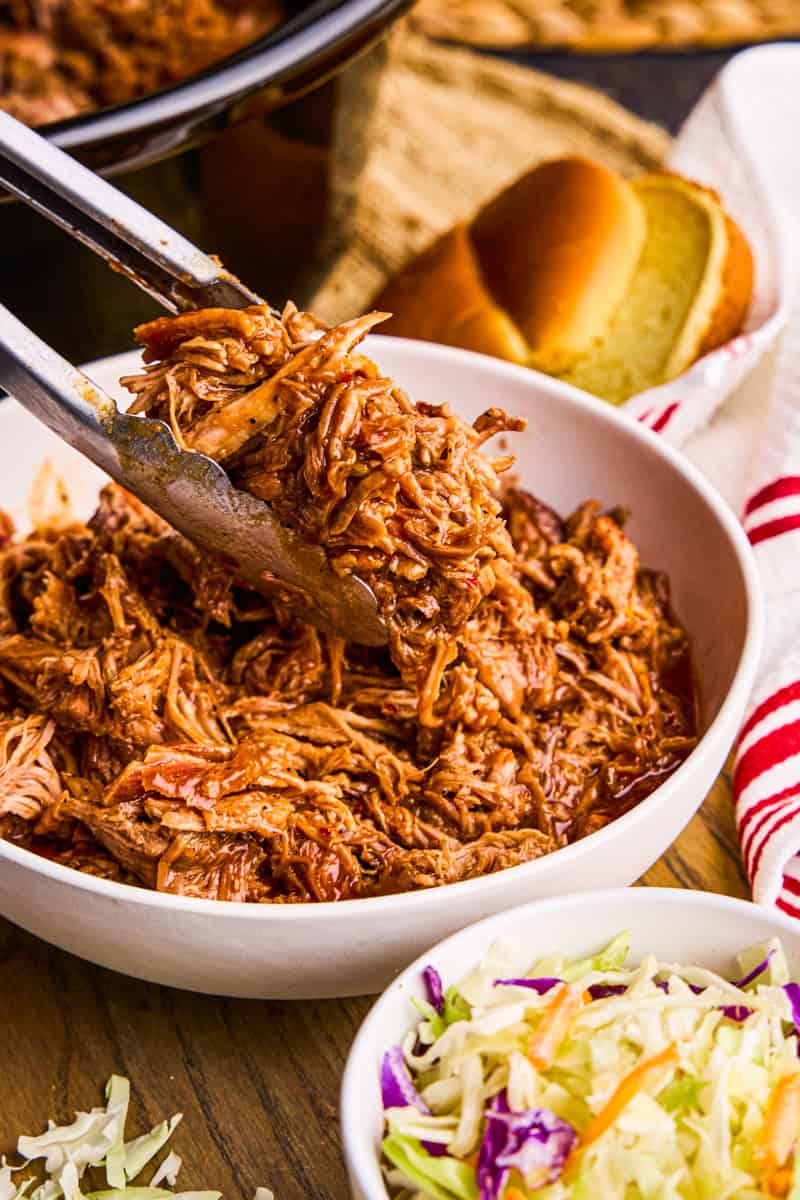 Tongs holding Slow Cooker BBQ Pulled Pork above a bowl.