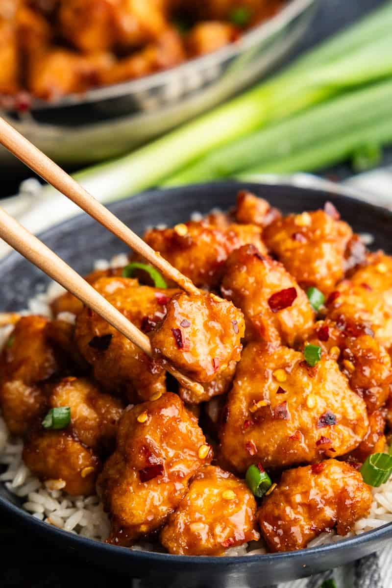 Closeup of General Tso Chicken on a plate of rice being lifted by chopsticks.