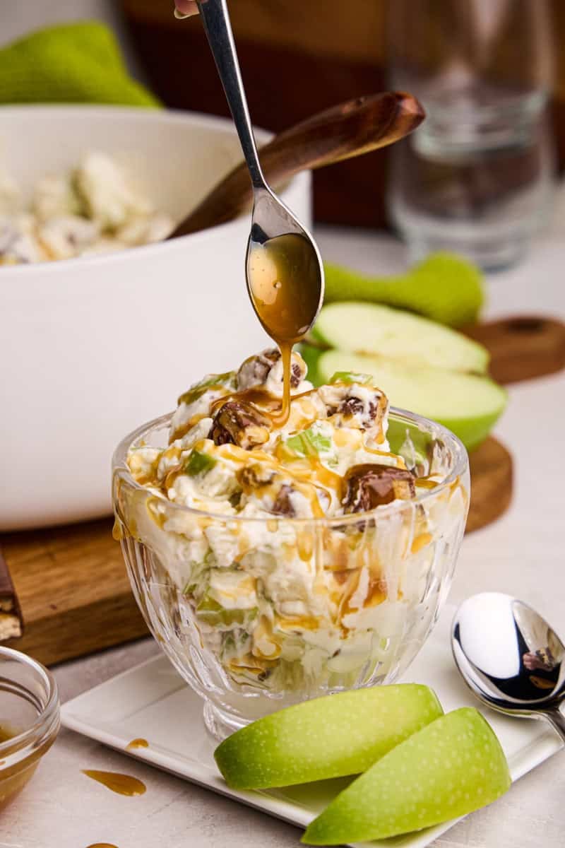 Apple Snickers Salad in a glass cup with a spoon drizzling caramel on top.