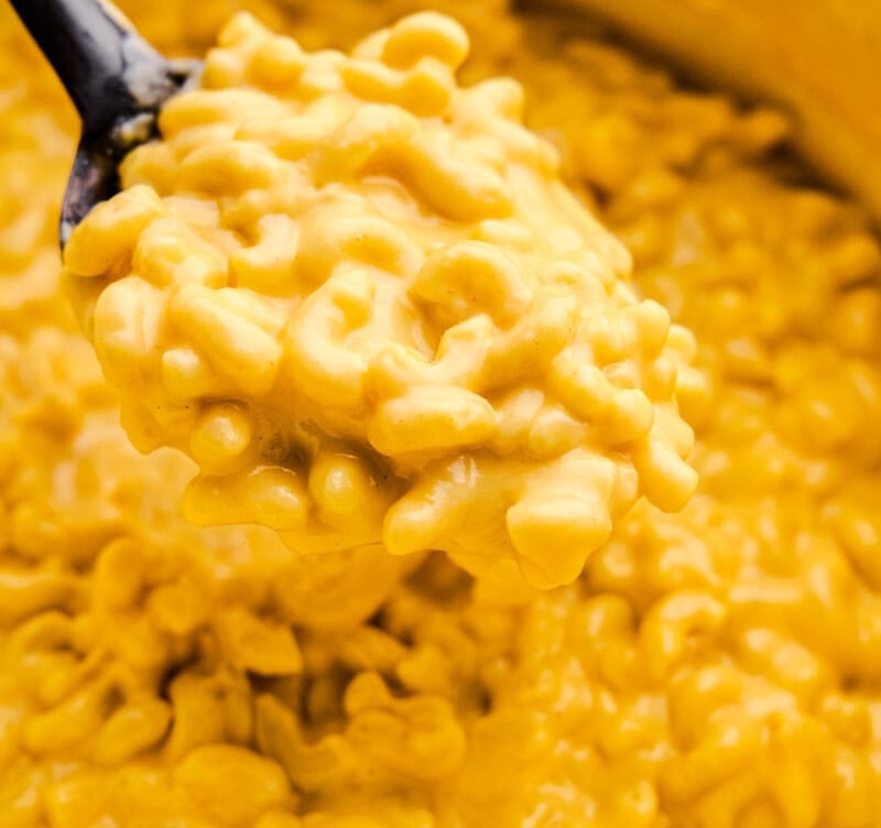 A serving spoon loaded with Easy Stovetop Mac and Cheese hovering over a cast iron pot full of Mac and Cheese.