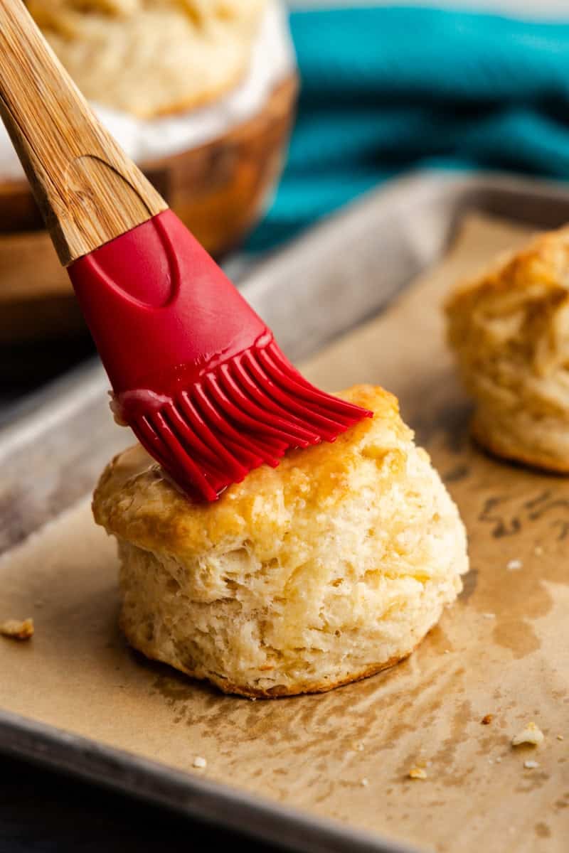 Closeup of butter being spread onto the top of a biscuit on a tray.