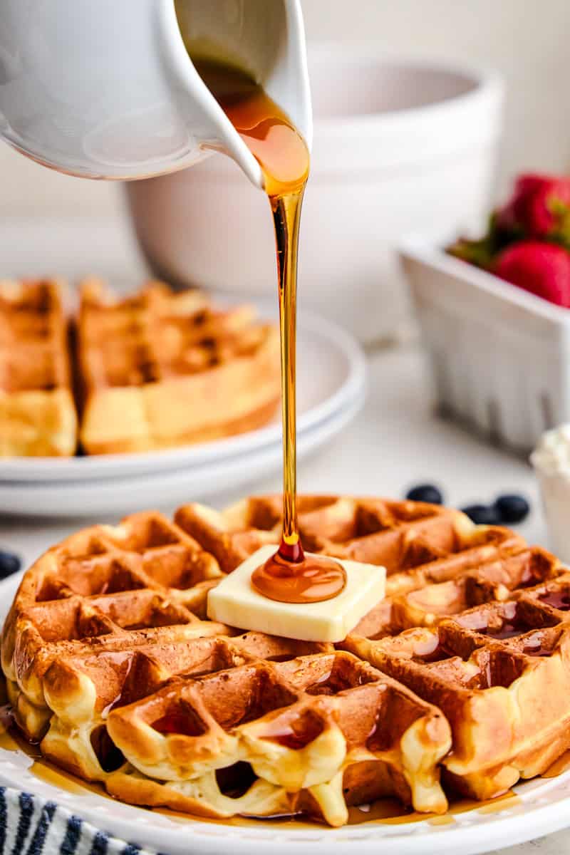 A stream of syrup pouring out and drizzling onto the center of a Greek Yogurt Waffle with a pat of butter.