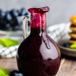 Pouring bottle of blueberry syrup with pancakes and blueberries in the background.