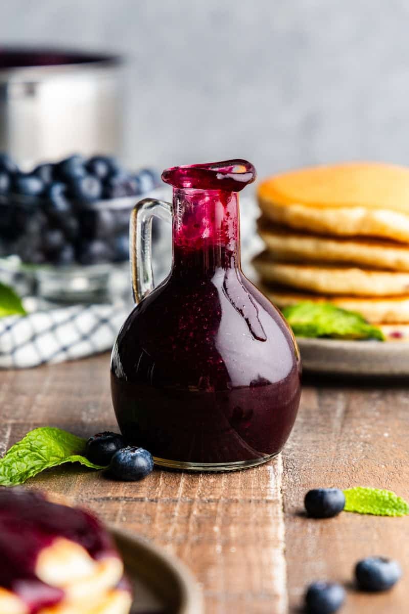 Pouring bottle of blueberry syrup with pancakes and blueberries in the background.