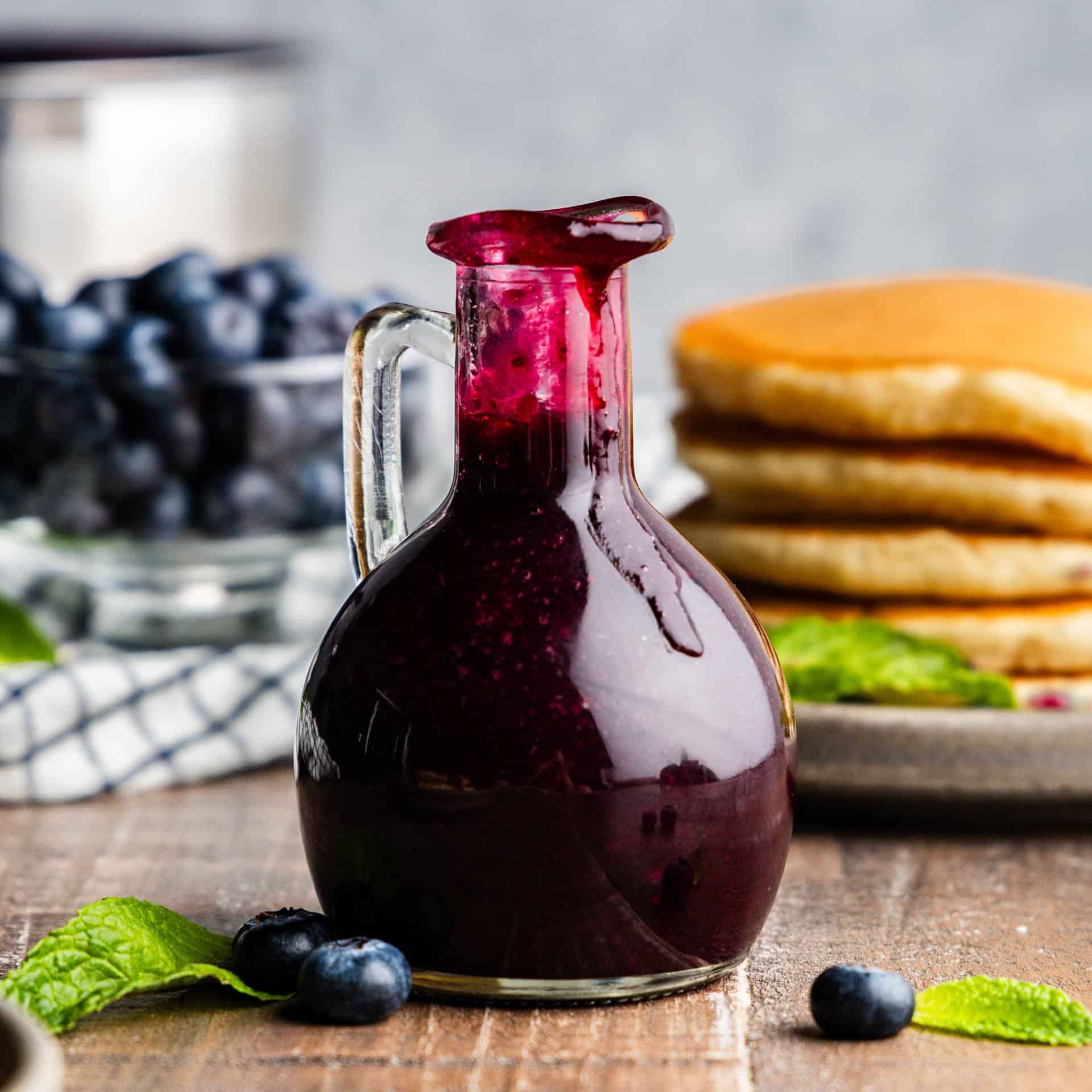 Pouring bottle of blueberry syrup with pancakes and blueberries in the background.