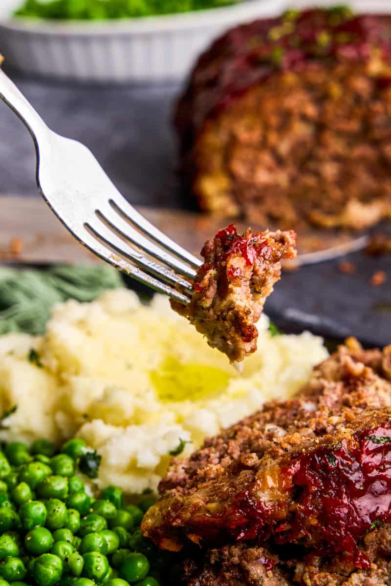 A bite of Momma's Meatloaf on a fork hovering over a plate with mashed potatoes and green peas.