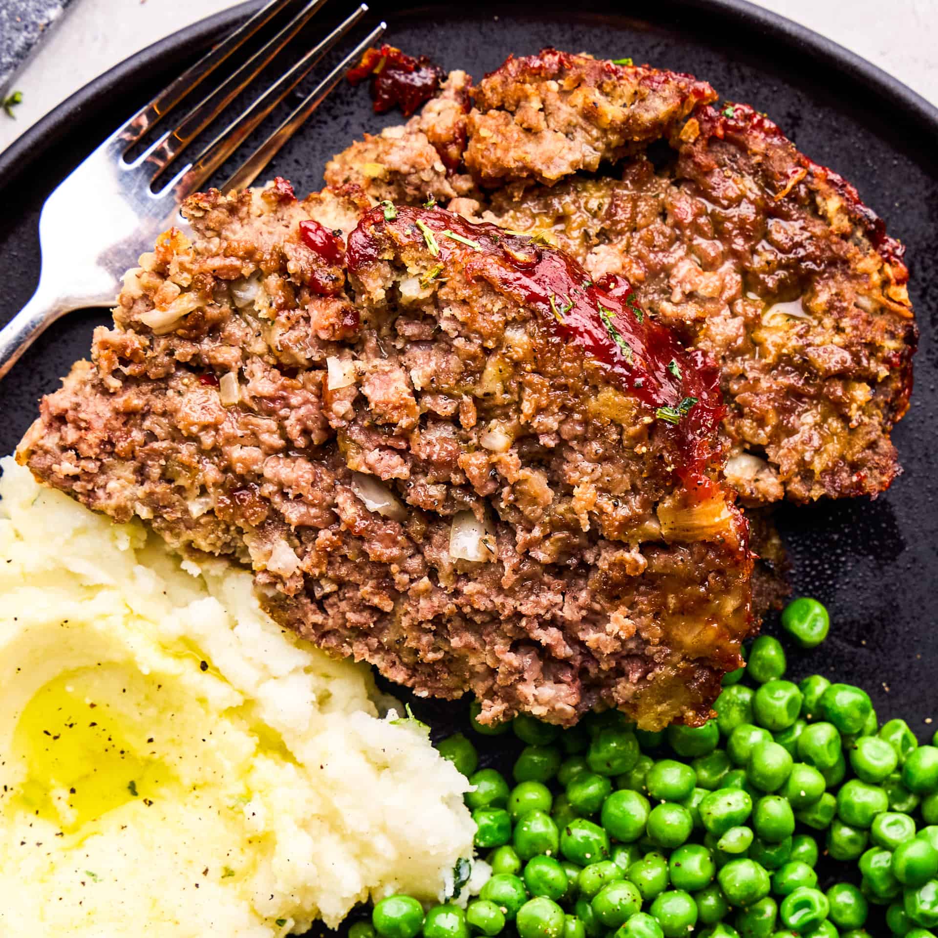 Two Slices of Momma's Meatloaf on a plate with mashed potatoes and green peas.