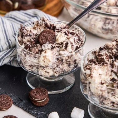 Oreo Fluff in a parfait cup with spoon in it and marshmallows and chocolate sandwich cookies scattered around.