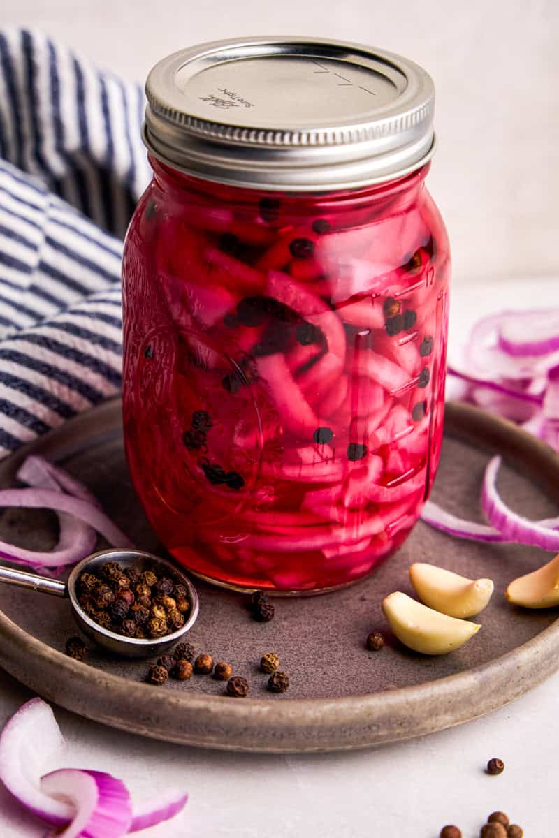 Pickled Red Onions in a glass jar with peppercorns and garlic ingredients next to it.