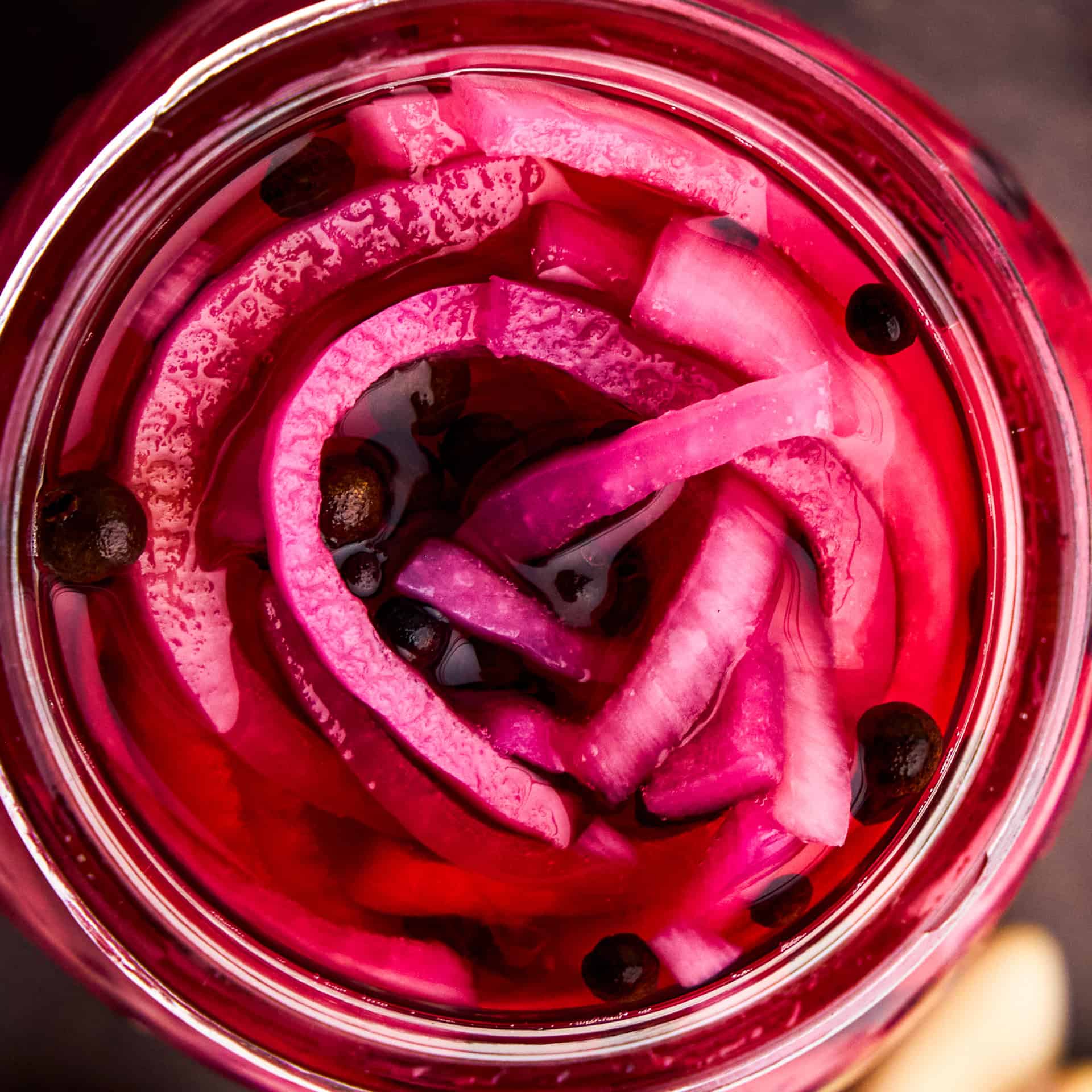 A closeup overhead image of Pickled Red Onions in a glass jar with peppercorns.