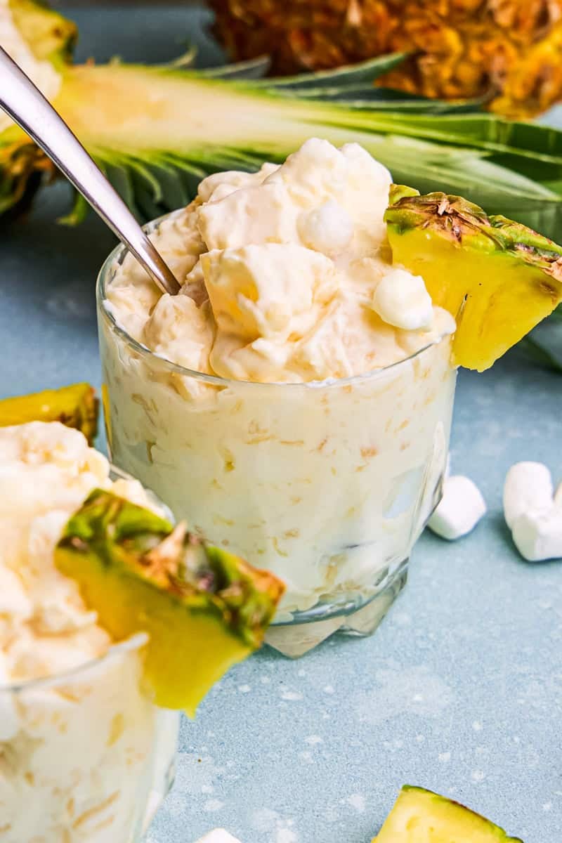 Pineapple Fluff in a glass cup with a spoon and surrounded by marshmallows and pineapple chunks.