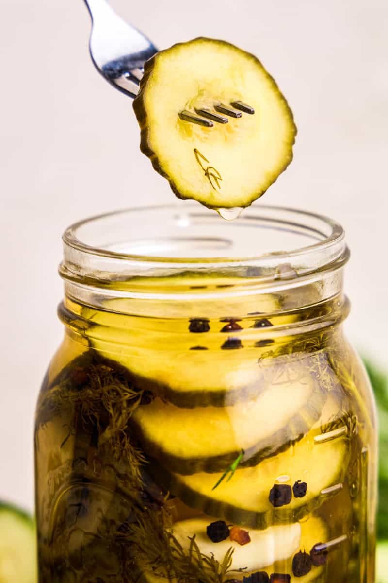 A pickle slice dripping in brine being held over the top of a glass jar of Refrigerator Pickles.
