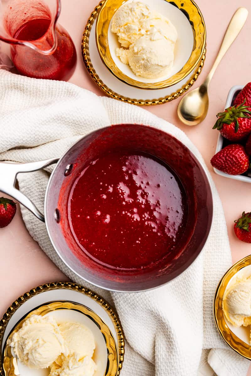 Strawberry syrup in a pot surrounded by bowls of ice cream and strawberries.