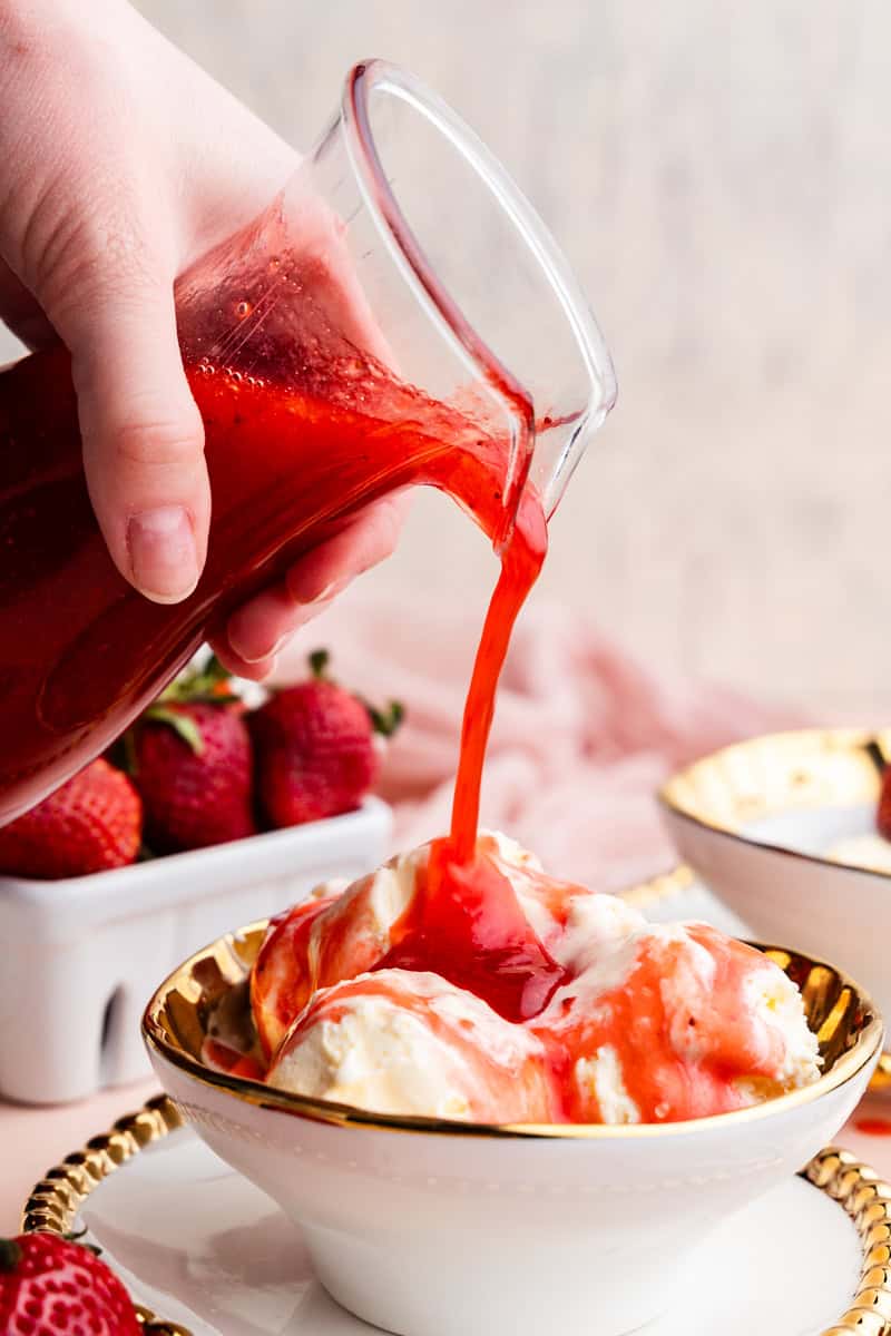 View of a pitcher of strawberry syrup being poured on bowl of ice cream.