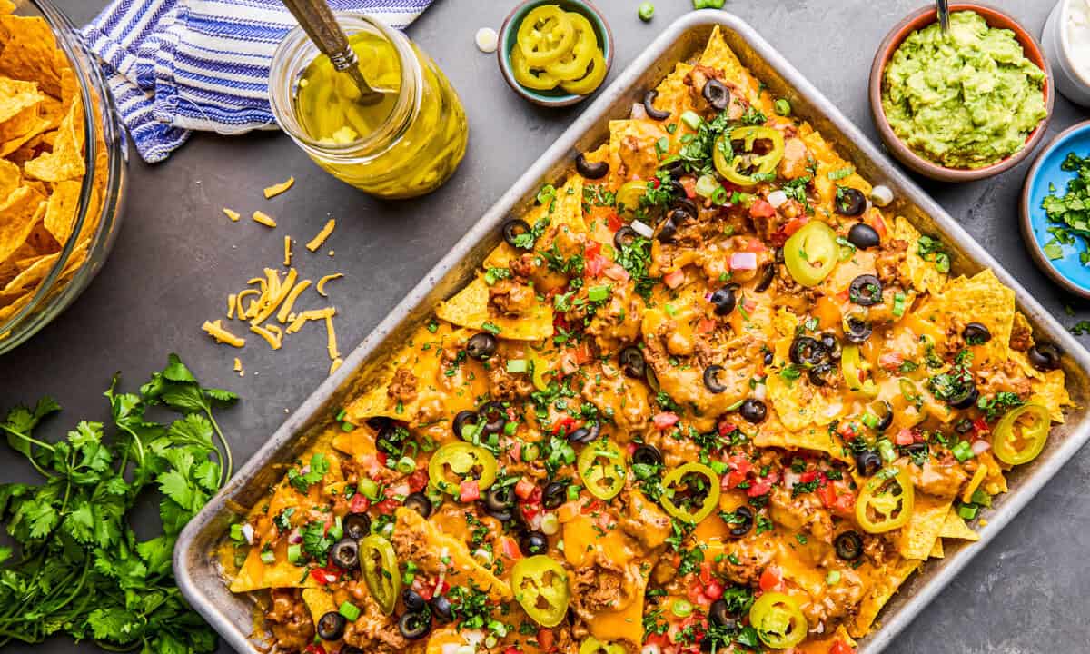 Overhead image of a pan of Ultimate Nacho Recipe with ingredients and toppings surrounding it.