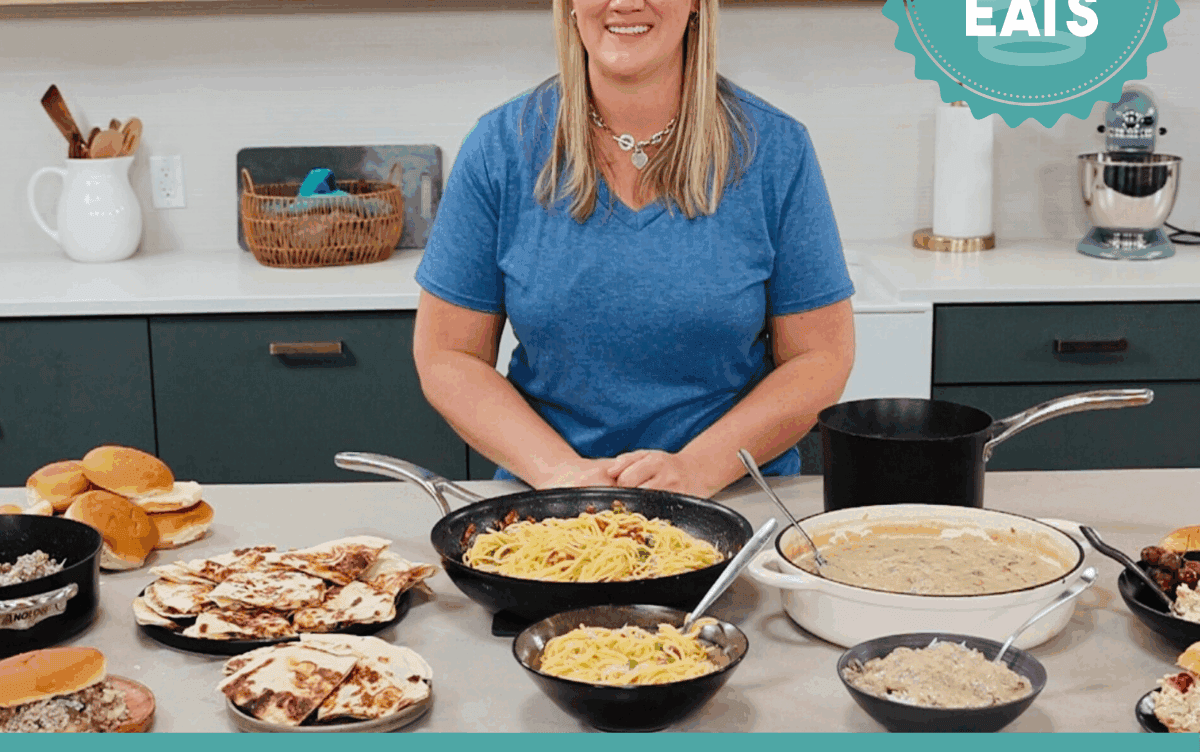A photo of Chef Cayt in The Stay At Home Chef Kitchen with the meals from Week 1 of the Cheap Eats Meal plan on display on the kitchen counter.