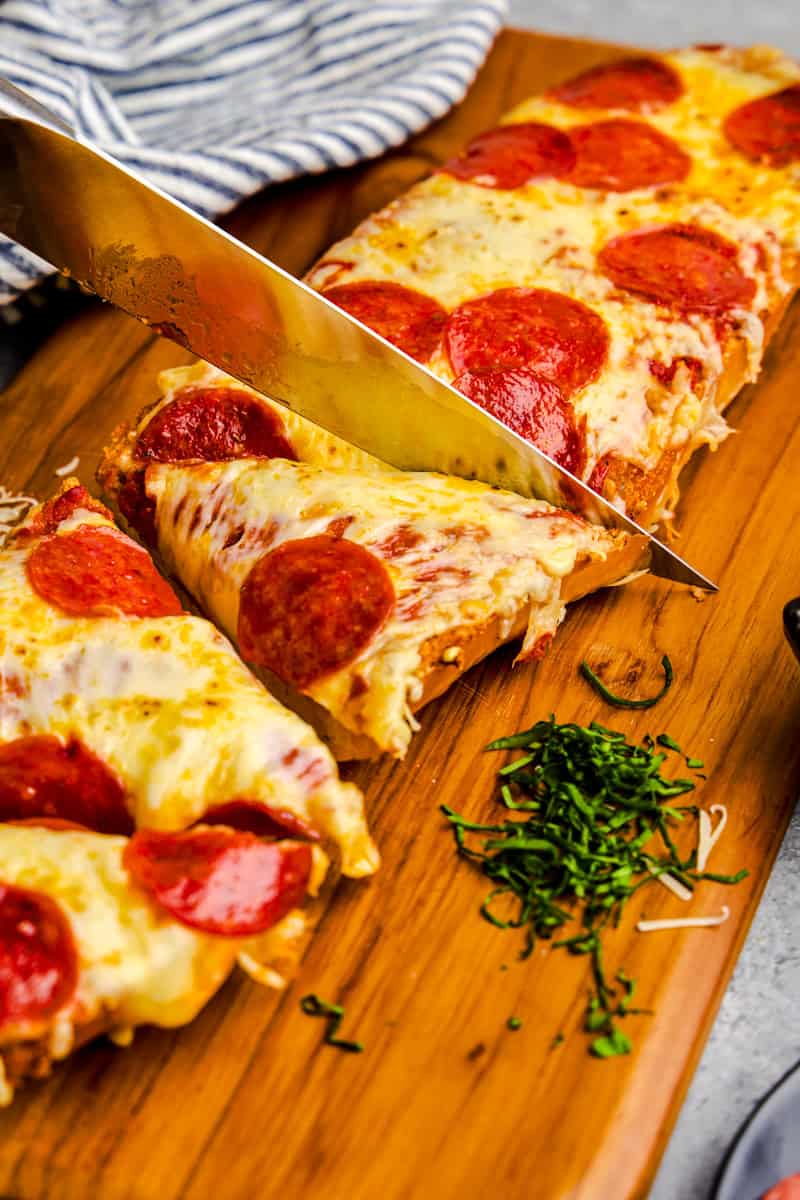 A knife cutting into French Bread Pizza on a wooden cutting board.