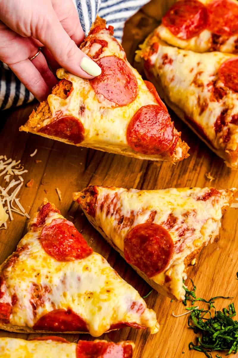 A hand grabbing a slice of French Bread Pizza from a cutting board with pepperonis, cheese and basil.