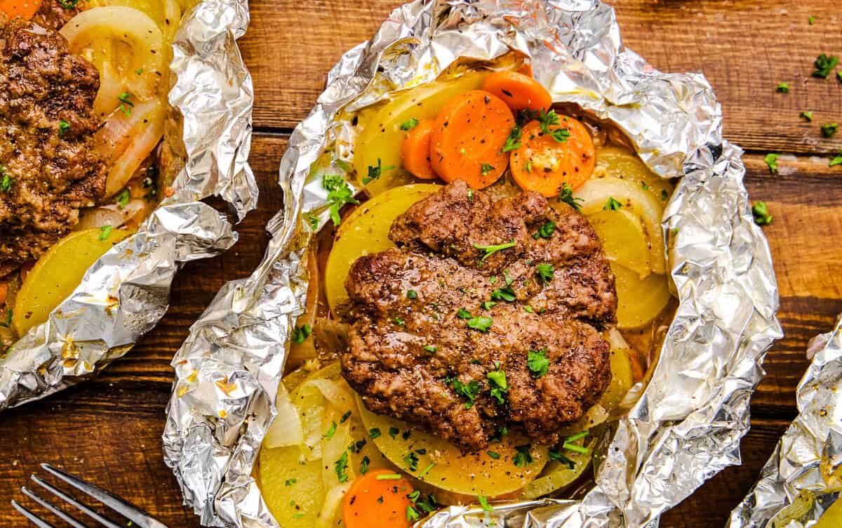 Open Hobo Foil Packet with burger, carrots, onions and potatoes on a wooden table.