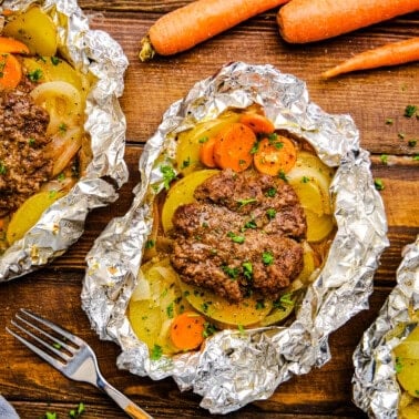 Open Hobo Foil Packet with burger, carrots, onions and potatoes on a wooden table.