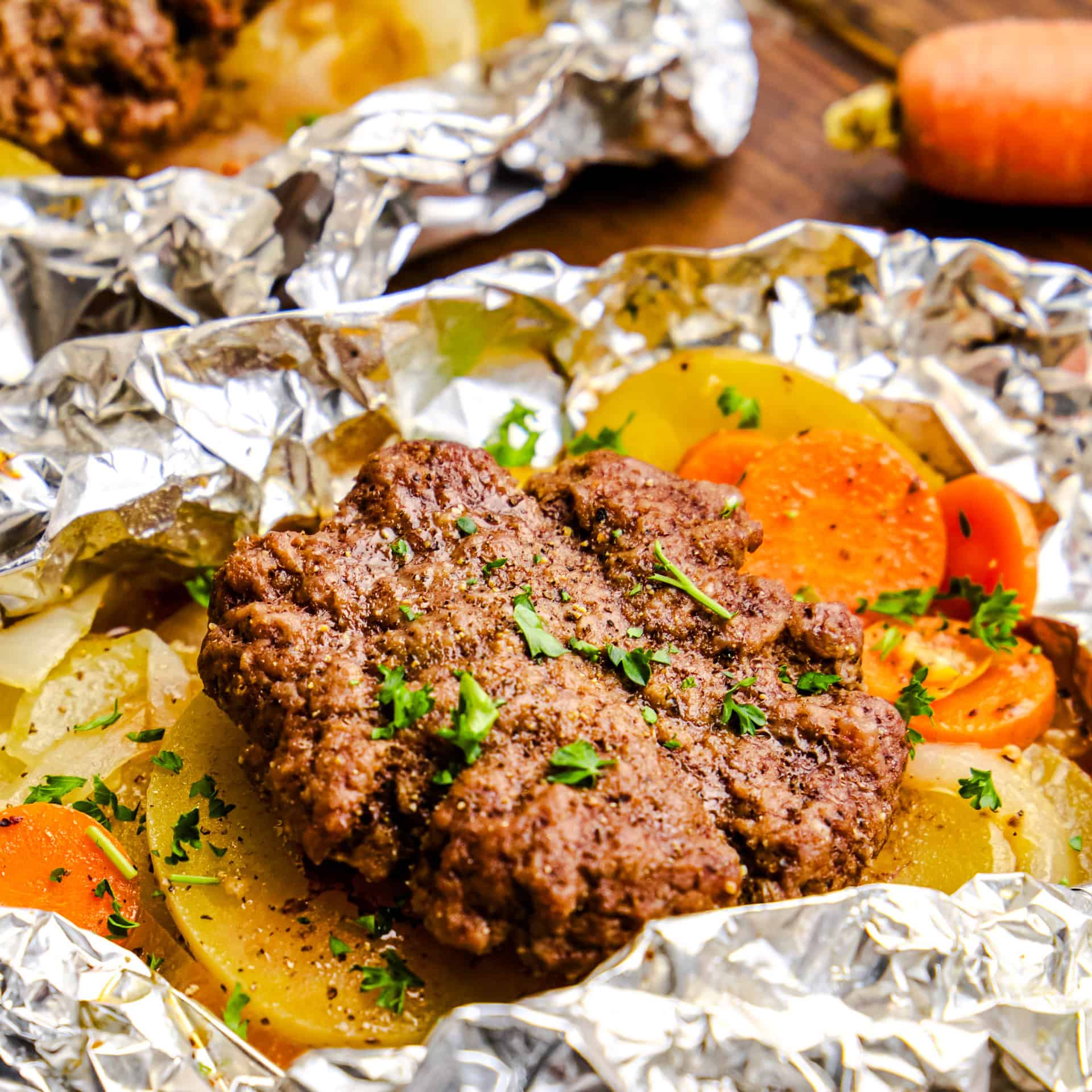 Open Hobo Foil Packet with burger, carrots, onions and potatoes on a wooden table.