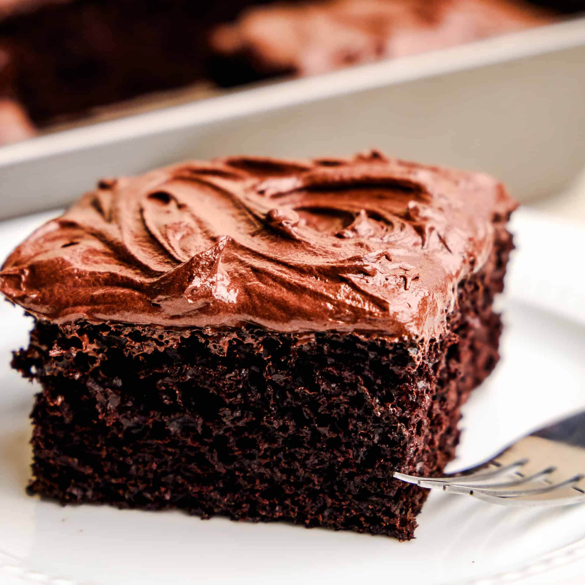A closeup image of a slice of Easy Chocolate Cake on a white plate with the cake pan in the background.