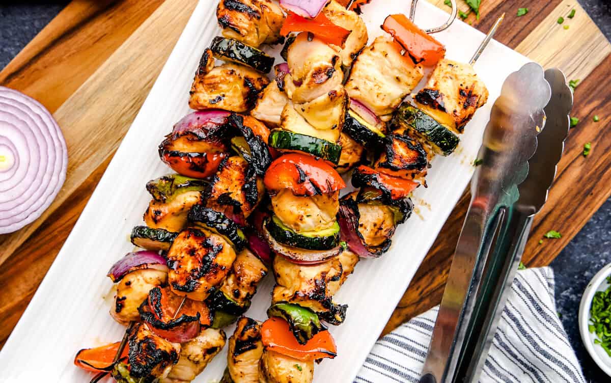 Juicy Marinated Grilled Chicken Kabobs on a white plate with tongs.