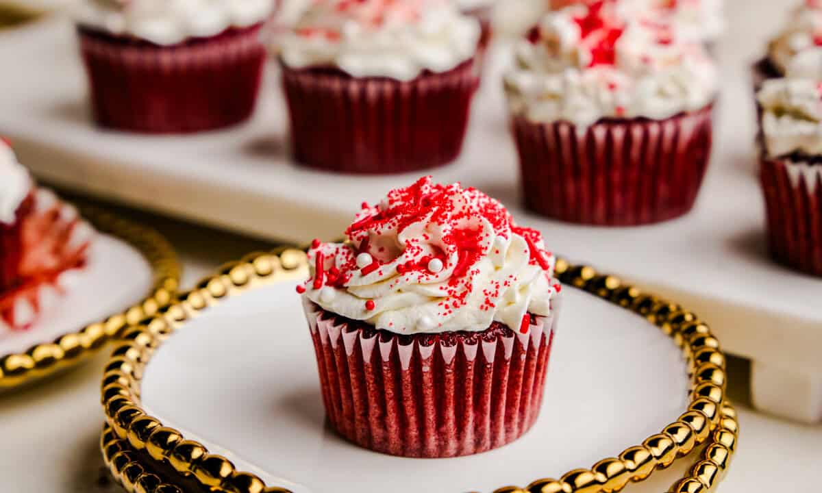 A single Most Amazing Red Velvet Cupcake on a fancy plate with gold trim in front of tray of cupcakes.