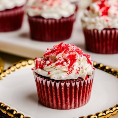 A single Most Amazing Red Velvet Cupcake on a fancy plate with gold trim in front of tray of cupcakes.