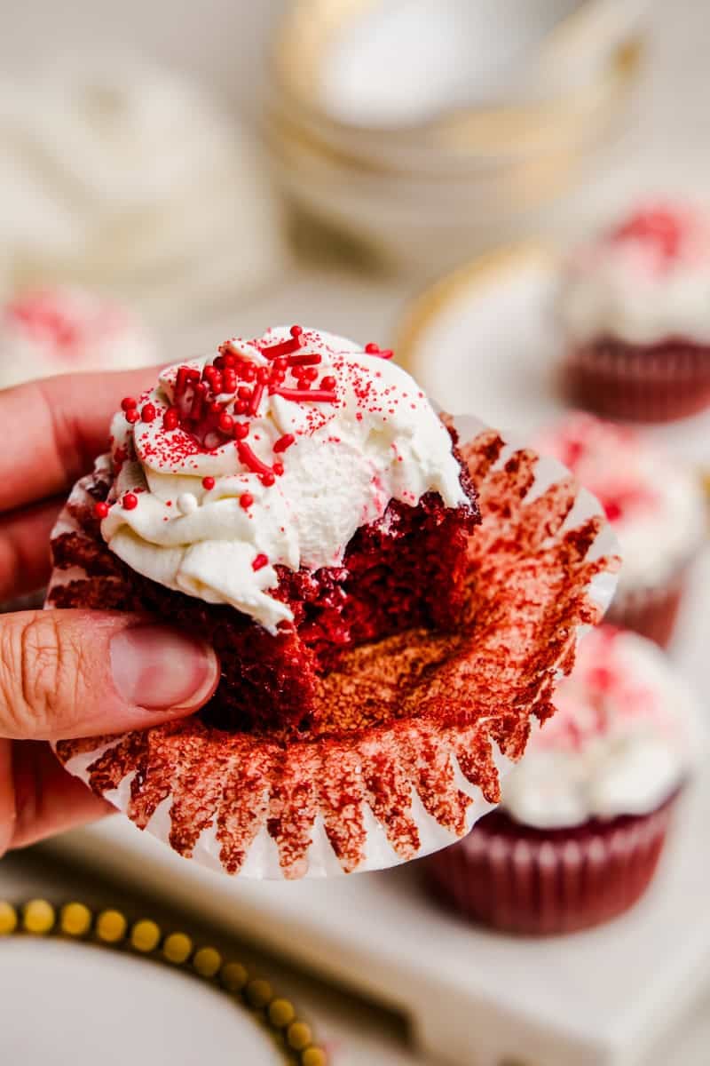 A hand holding a Most Amazing Red Velvet Cupcake topped with fluffy white frosting with a bite out of it.