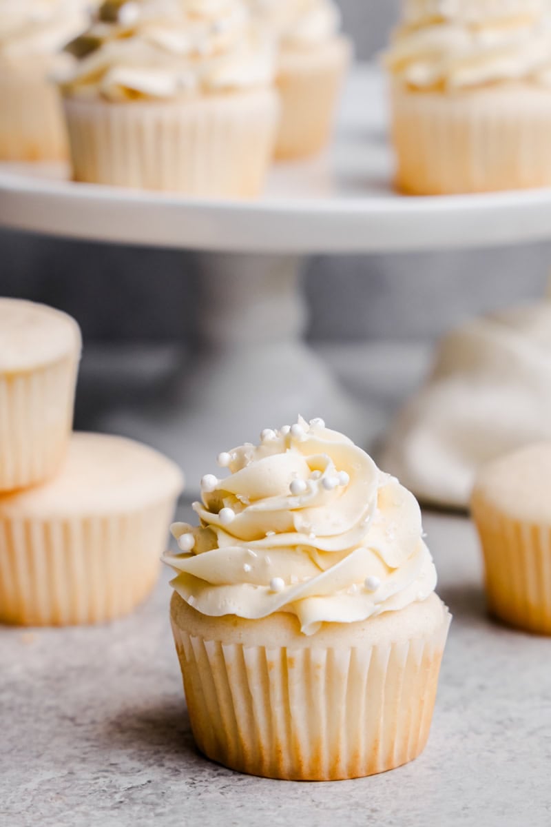 A Most Amazing White Cupcakes with crystal sugar and white sprinkles in front of a cake stand full of cupcakes.