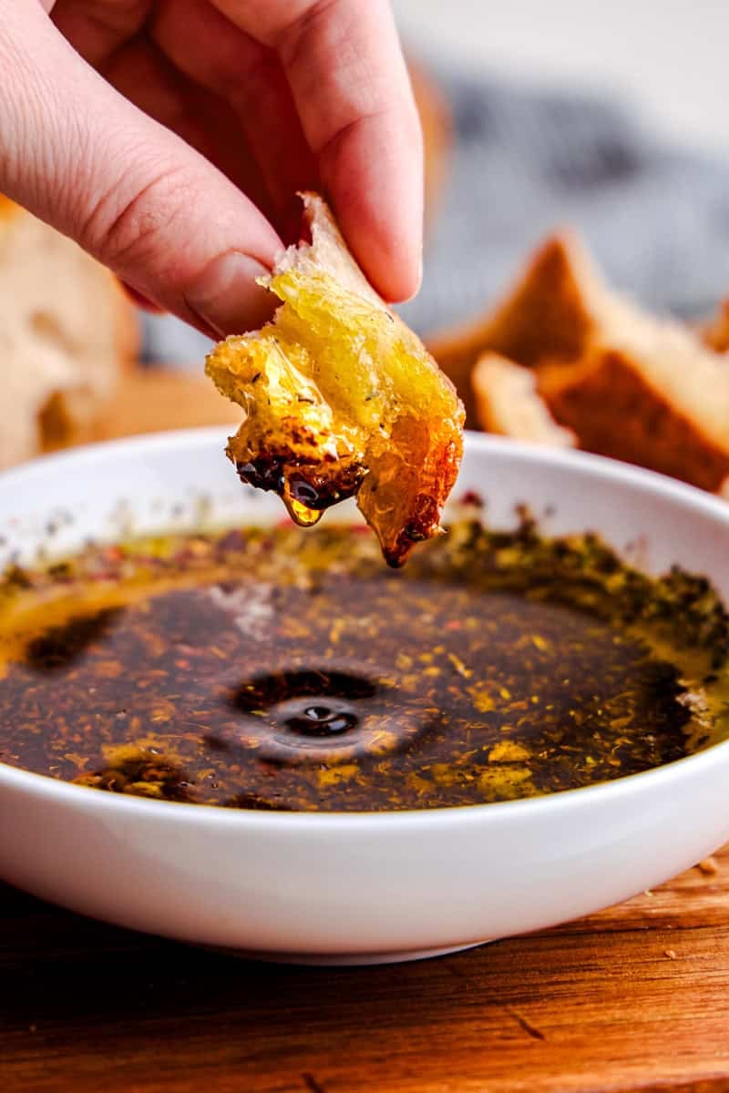 A hand dipping a piece of bread into a bowl of Olive Oil Bread Dip on a cutting board.