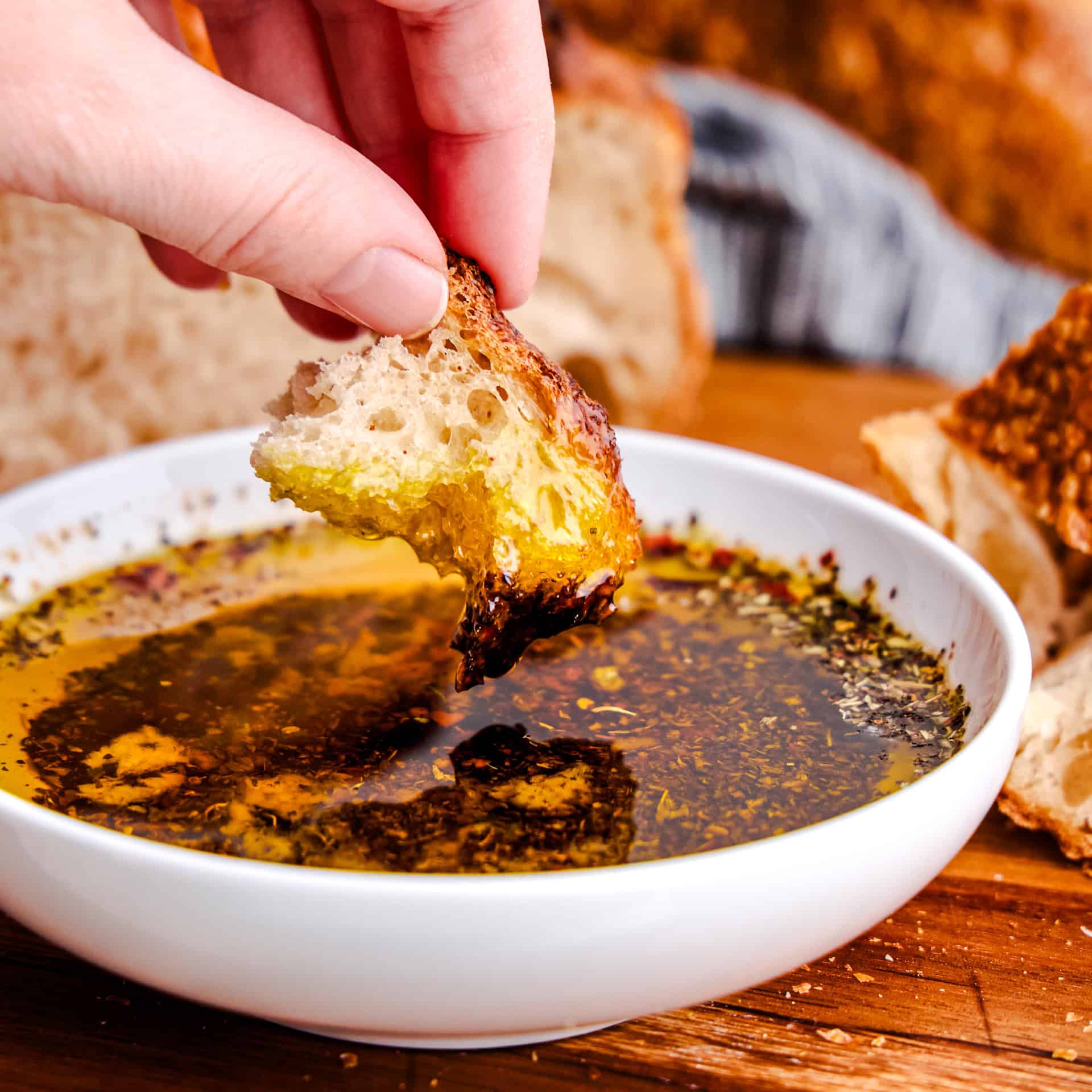A hand dipping a piece of bread into a bowl of Olive Oil Bread Dip on a cutting board.
