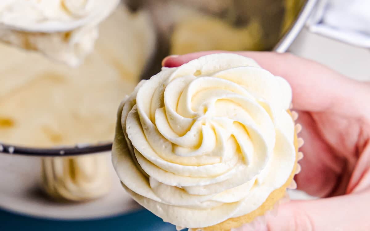 Perfect Cream Cheese Frosting swirled onto a cupcake with a mixing bowl full of frosting in the background.